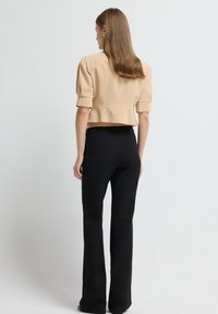 Vrouw met steil bruin haar, gekleed in een beige top met korte mouwen en hoge zwarte broeken, staande voor een effen witte achtergrond.