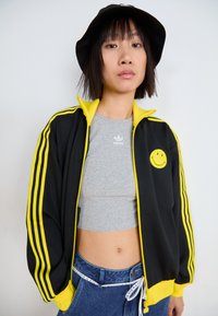 Černá zipová bunda se žlutými pruhy a emblemem usmívající se tváře, nošená přes šedý crop top. Kombinováno s modrými džíny s vysokým pasem.