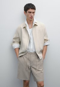 Lys beige linnedjakke over en hvid skjorte, parret med lyse khaki shorts. Casual snit med synlige sømdetaljer og snoreliv.