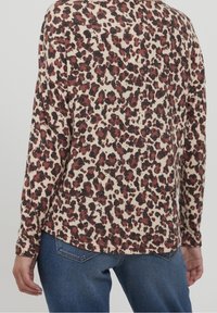 T-shirt à manches longues beige avec un motif léopard brun et noir ; tissu doux, ourlet arrondi et coupe légèrement ample. Porté avec un jean bleu.