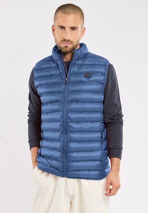 Homme portant une veste sans manches matelassée bleue sur une chemise noire à manches longues et un pantalon de couleur claire, debout les mains dans les poches.