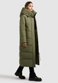 Garšs olīvu zaļš pūkains mētelis ar augstu apkakli, kuram ir quiltētas daļas un sānu kabatas, kas saskaņots ar melniem biksēm un masīviem melniem zābakiem.