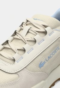 Μπεζ αθλητικά παπούτσια με υλικά από δίχτυ και σουέντ, με ανοιχτό μπλε λεπτομέρειες, επίπεδα κορδόνια και λαστιχένια σόλα. Λογότυπο Lacoste στο πλάι.