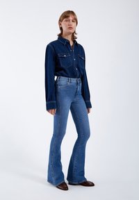 Camisa de mezclilla en azul oscuro con dos bolsillos en el pecho; jeans azules de tiro alto y acampanados; textura suave y corte clásico.