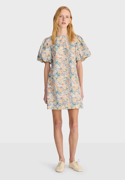 Femme debout portant une robe courte à imprimé floral avec des manches bouffantes, associée à des chaussures lacées couleur crème sur un fond clair uni.