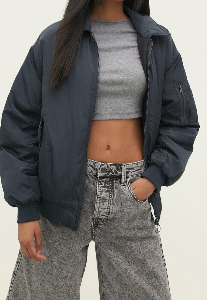 Marineblauwe bomberjack met elastische manchetten en een ritszak, gedragen over een grijs cropped topje. Gecombineerd met lichtgrijze, high-waisted denim jeans.