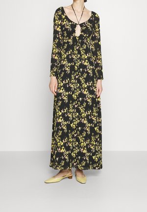 Vrouw draagt een zwarte maxi-jurk met lange mouwen met gele en roze bloemenpatronen en gele slingback schoenen met lage hak.