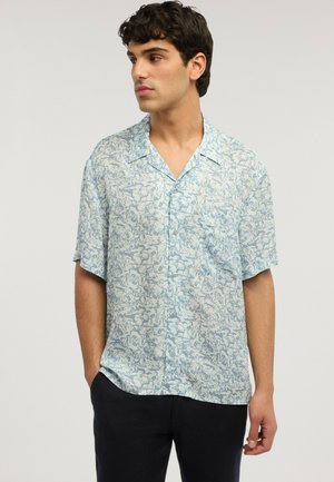 Mann trägt ein kurzärmliges hellblaues Hemd mit abstraktem Blumenmuster, schwarze Hose, Hände in den Taschen, schaut nach links.