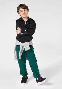 Sweatshirt preta com fecho de correr e logótipo "Champion", cinto cinzento na cintura, calças cargo verdes e ténis pretos com detalhes coloridos. Textura lisa.