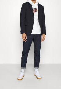 Blazer negro sobre una sudadera blanca con un logo, combinado con jeans oscuros de mezclilla enrollados y zapatillas blancas con suelas de goma, de pie sobre un fondo simple.