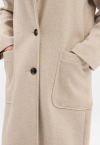 Beige Wollmantel mit Knopfverschluss, zwei Fronttaschen und weicher Textur. Das Design umfasst umrandete Taschendetails.