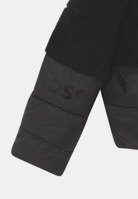 Černá fleecová bunda s texturovanou horní částí a hladkým spodním rukávem z materiálu puffer. Obsahuje embosované logo "BOSS" ve tmavém, reflexním materiálu.