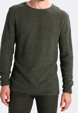 Muž stojící proti jednoduchému světlemu pozadí, má na sobě tmavě zelený texturovaný svetr s dlouhým rukávem a tmavé kalhoty ladící s svetrem.