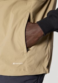 Giacca beige chiaro con tasca laterale, dotata di un polsino a coste nero. Il tessuto appare liscio e leggero, con un piccolo logo.