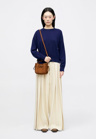 Granatowy sweter z dzianiny, beżowe spodnie w kant i brązowa torba zamszowa z skórzanym paskiem. Model stoi na jednolitym tle.