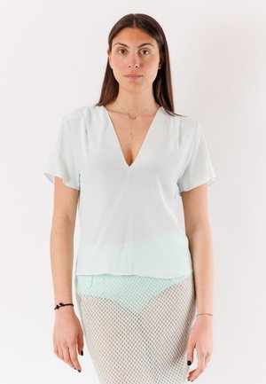 Donna con capelli lunghi e scuri che indossa una maglietta azzurro chiaro con scollo a V e maniche corte e una gonna bianca a rete, in piedi davanti a uno sfondo semplice.