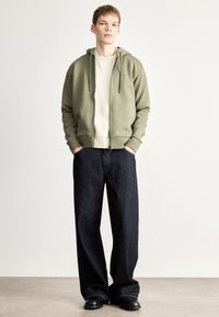 Felpa con cappuccio verde oliva con zip, abbinata a un maglione a coste beige chiaro e jeans larghi in denim scuro, completata da calzature nere. Design minimalista.