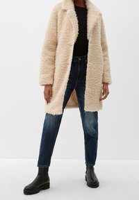 Manteau en fausse fourrure crème avec un col châle, porté sur un haut en tricot noir et un jean droit bleu. Des bottes noires à talons complètent l'ensemble.