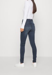 Jeans skinny azul oscuro de denim con cintura alta. Presenta dos bolsillos traseros y costuras laterales verticales, combinados con zapatillas blancas.