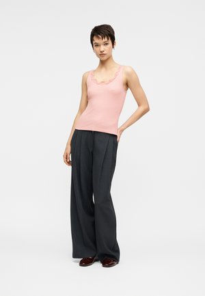 Lühikeste tumedate juustega naine, kes kannab roosat varrukateta topi pitsist äärega, laiu tumedaid halli värvi pükse ja tumedaid poleeritud jalanõusid, taust on ühevärviline.