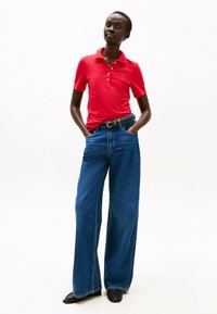 Polo rosso con colletto e bottoni abbinato a jeans in denim blu a gamba larga. Il modello indossa una cintura nera e scarpe nere. Sfondo bianco uniforme.