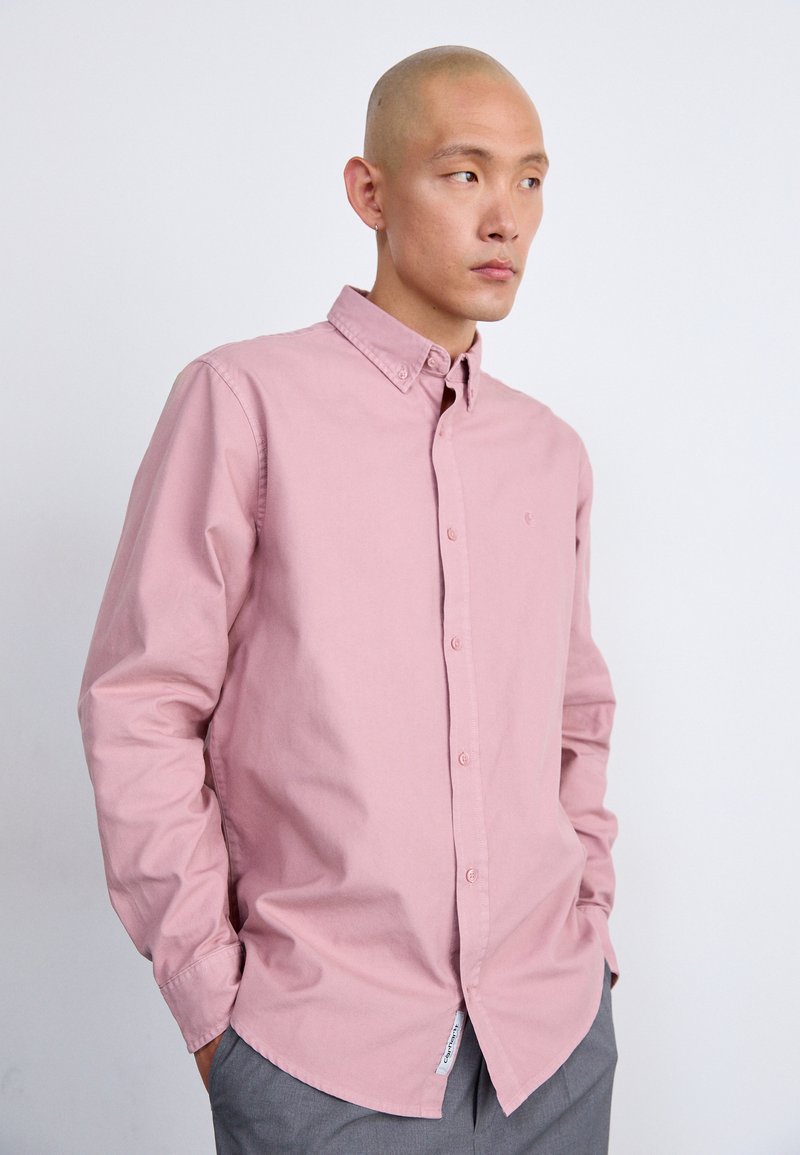 Camicia button-up rosa chiaro con colletto, maniche lunghe e logo a patch sul petto, abbinata a pantaloni grigi. Tessuto di cotone liscio.