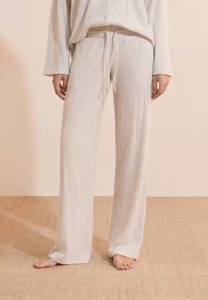 Personne portant un pantalon de détente beige ample côtelé avec cordon, debout pieds nus sur un sol beige texturé devant un mur beige uni.