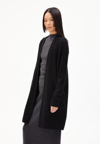 Schwarzer Cardigan aus einem weichen Strickmaterial, mit langen Ärmeln, offenem Front und seitlichen Taschen, über einem grauen gerippten Pullover getragen.