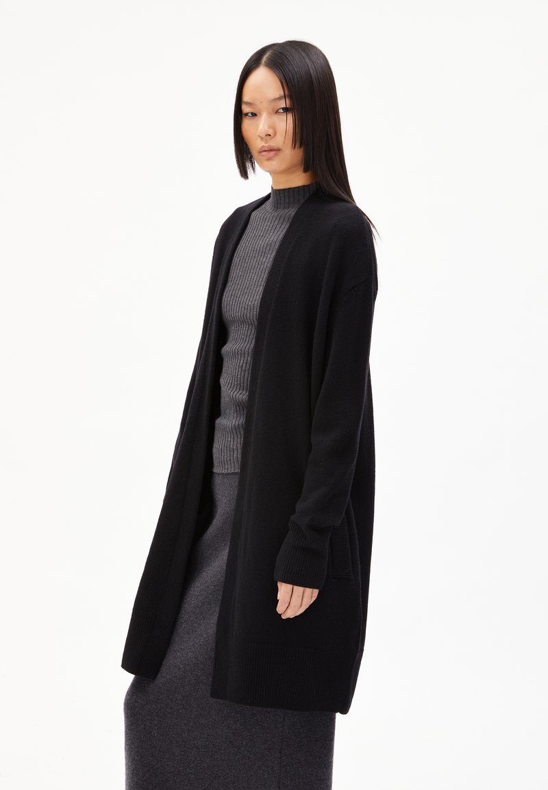 Schwarzer Cardigan aus einem weichen Strickmaterial, mit langen Ärmeln, offenem Front und seitlichen Taschen, über einem grauen gerippten Pullover getragen.