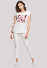 Junge Frau in einem weißen T-Shirt mit roten und schwarzen Herzmustern, weißen Hosen und weißen Sneakers, die vor einem schlichten Hintergrund steht.