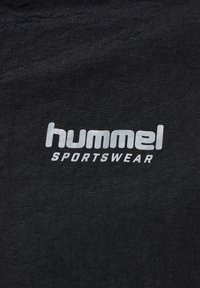 Svart tyg med en framträdande, reflektiv silverlogo som står "hummel SPORTSWEAR" i en djärv, modern stil.