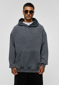 Grijze fleece hoodie met een voorzak en een platte trekkoordkap. Textuurstof, losse pasvorm en ribgebreide manchetten. Model draagt ronde zonnebril.