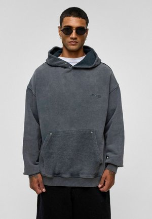 Grijze fleece hoodie met een voorzak en een platte trekkoordkap. Textuurstof, losse pasvorm en ribgebreide manchetten. Model draagt ronde zonnebril.
