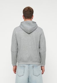 Mężczyzna tyłem, ubrany w jasnoszary hoodie i jasnoniebieskie dżinsy, na jednolitym białym tle.