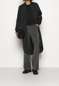 Černý prošívaný dlouhý kabát s vroubkovanými manžetami, v kombinaci se šedými kalhotami s širokými nohavicemi a černými platformovými botami. Drží černou kabelku.