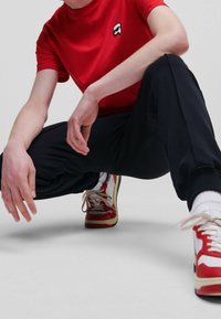 Rød bomulds t-shirt med et sort og hvidt logo, kombineret med sorte bukser og sneakers i rød, hvid og gul. Modellen knæler med hænderne på knæene.