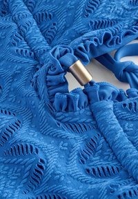 Blauwe textured stof met bloemenborduursel, met gerimpelde randen en een bronzen accent. Gladde oppervlakte met ingewikkelde patroondetails.