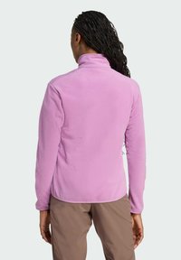 Rosa Fleecejacke mit hohem Kragen und langen Ärmeln, die eine glatte Textur und ein tailliertes Design aufweist, getragen über beigen Hosen.