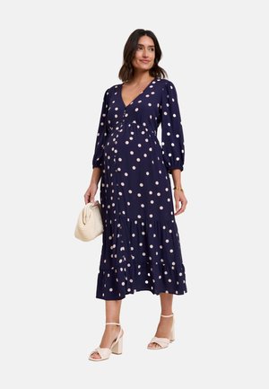 Zwangere vrouw draagt een marineblauwe midi-jurk met polkadots en lange mouwen, houdt een beige gevlochten handtas vast en draagt beige blokhakken.