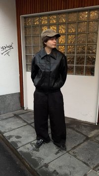 Egy személy fekete bőrkabátot, bő fekete nadrágot és barna sapkát visel, és egy nedves járdán áll egy üvegtéglás ablak előtt.