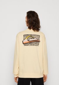 Sudadera de color beige claro con mangas largas, que presenta un diseño gráfico en la espalda en tonos negro, naranja y amarillo. Textura suave, corte holgado.