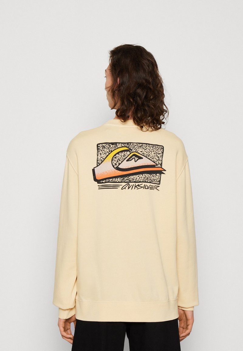 Sudadera de color beige claro con mangas largas, que presenta un diseño gráfico en la espalda en tonos negro, naranja y amarillo. Textura suave, corte holgado.