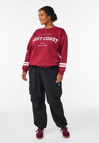 Bordeaux sweatshirt med hvid "WEST COAST" tekst, hvide striber på ærmerne; sorte cargo bukser; bordeaux sneakers med lyse såler.