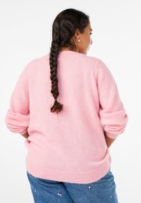 Rožnat, mehak pulover z ohlapnim krojem in zavihanimi rokavi, izdelan iz puhastega materiala. Nošen z jeansa z majhnimi cvetličnimi poudarki.