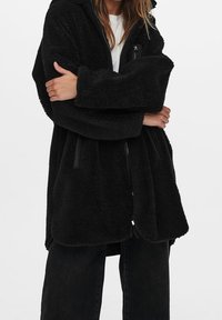 Chaqueta de forro polar negra con cierre de cremallera, ajuste holgado, dobladillo redondeado y dos bolsillos frontales con cremallera. Tejido texturizado con una apariencia suave.