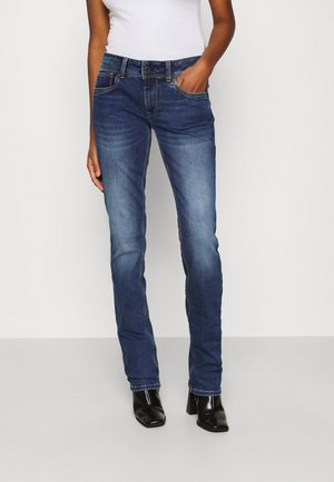 Osoba v modrých džínech s úzkým střihem a černých kotníkových botách na podpatku, stojící s jednou rukou v kapse před jednobarevným pozadím.