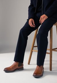 Brown leather Chelsea boots with elastic side panels, rounded toe, and textured finish, paired with dark pants and a wooden stool.