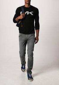 Pullover nero a maniche lunghe con logo grigio, jeans grigi e sneakers blu. L'outfit è completato da uno zaino scuro con un accento marrone.