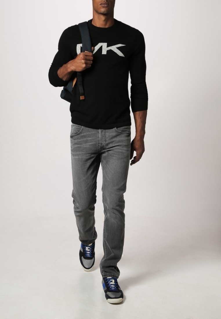 Pullover nero a maniche lunghe con logo grigio, jeans grigi e sneakers blu. L'outfit è completato da uno zaino scuro con un accento marrone.