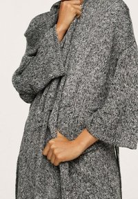 Cardigan gris texturé avec manches larges et fermeture à nouer. Le tissu semble doux avec un motif tacheté. Main visible tenant le devant.